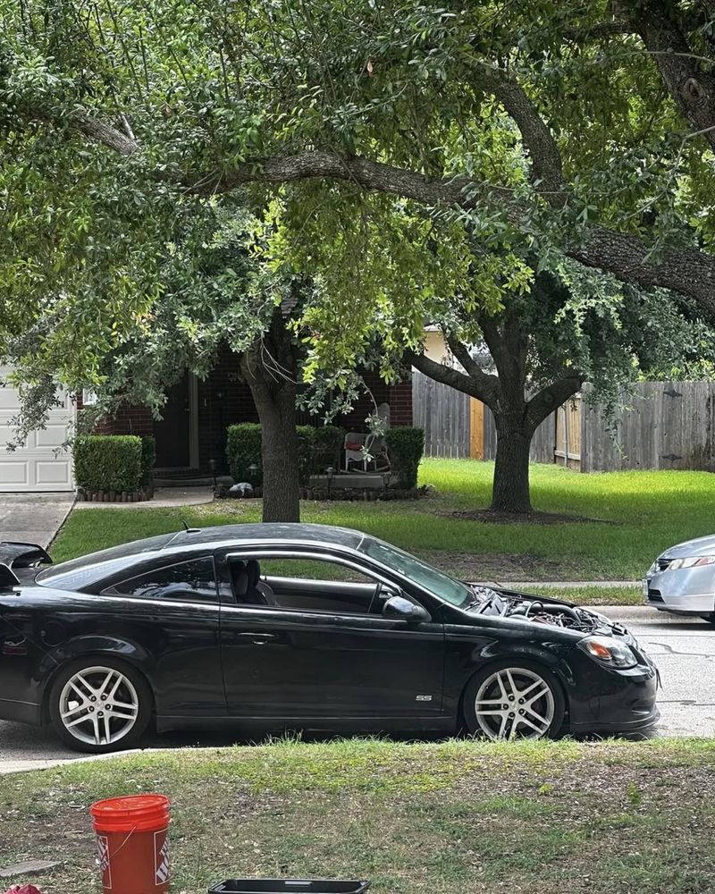 The black Cobalt SS coupe from the side, parked at the curb with the hood open
