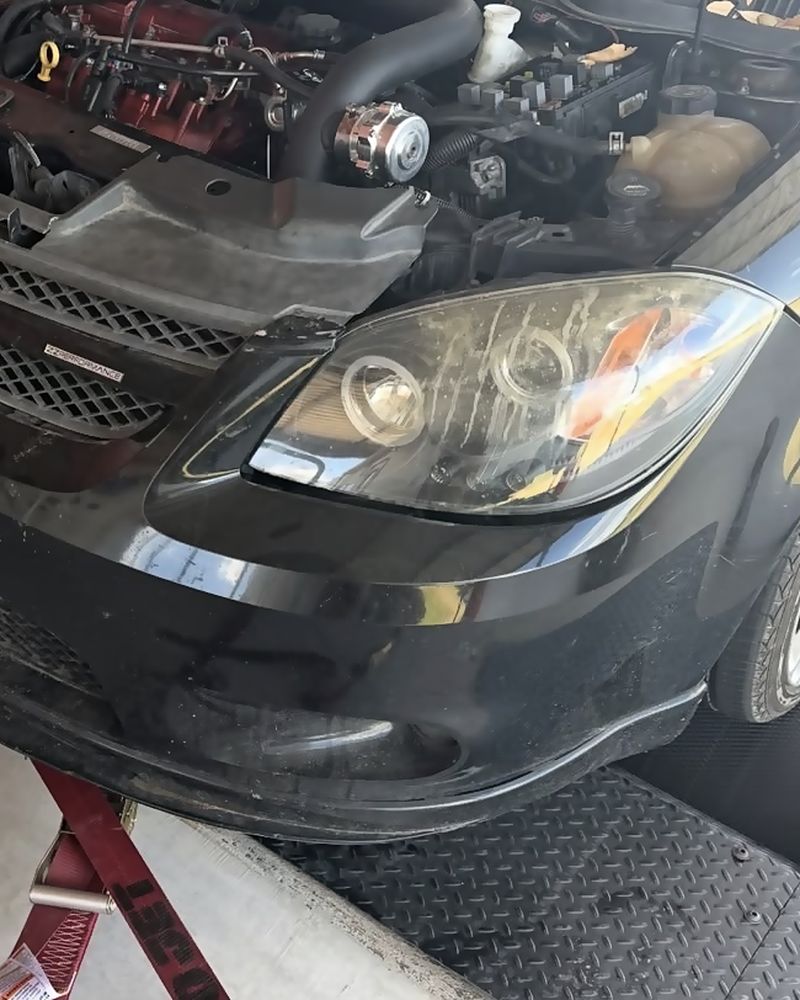 The front of the black Cobalt strapped to the dyno rollers with the hood open, turbo plumbing visible
