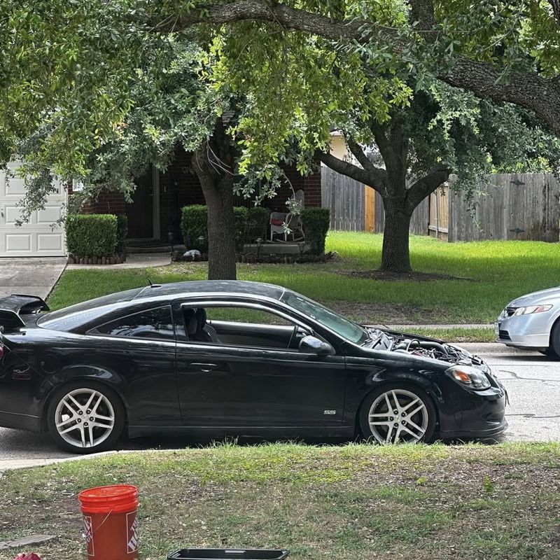 The black Cobalt SS coupe in profile, parked at a curb under oak trees with its hood raised