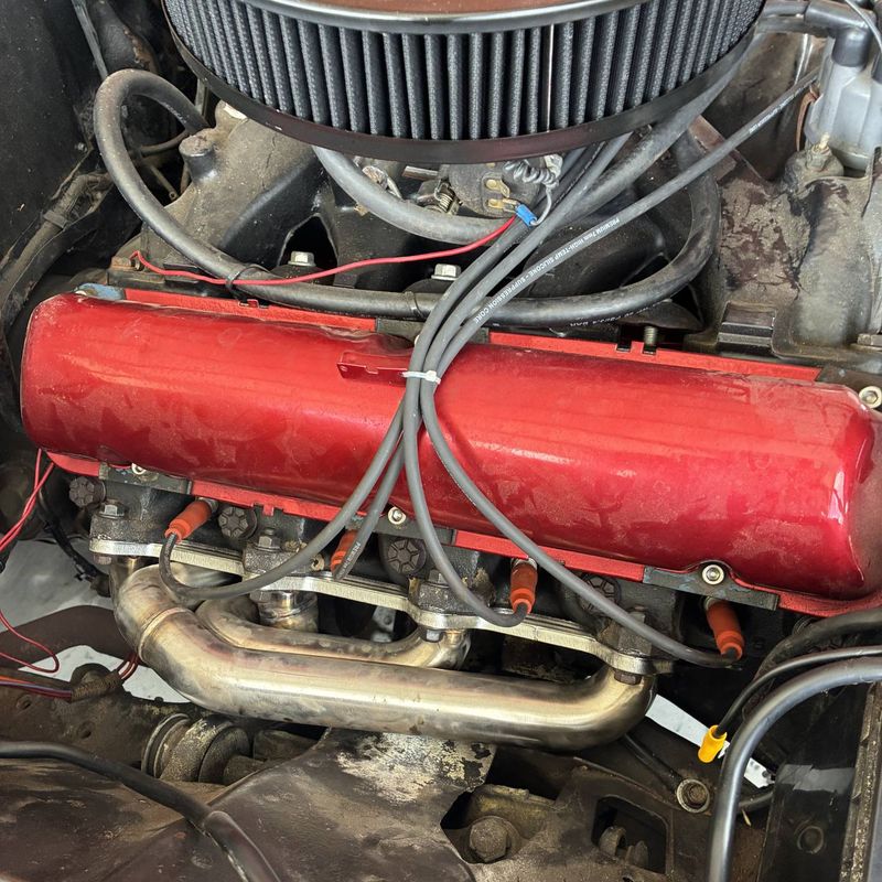 A carbureted V8 in the bay, chrome air cleaner above a red valve cover and stainless headers